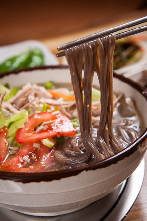Chopsticks picking up cold noodles with sliced carrots, cucumber, tomatoes and chicken meat, in china bowlの写真素材