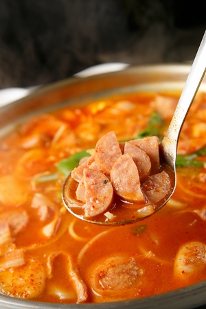 ladle scooping ham from Budae jjigae, spicy sausage stew with ingredients like ham, noodles, kimchi, rice cake, kimchi and garlic chives in fry panの写真素材