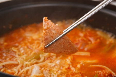 chopsticks holding ham from boiling Budae jjigae, spicy sausage stew with noodles,cheese, ham, pork, bean sprouts, garlic chives and onion on fry panの写真素材