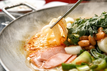 chopsticks holding udon noodles from Budae jjigae, spicy sausage stew with ingredients like onion,...