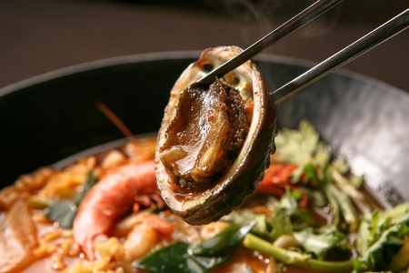 Chopsticks grabbing abalones from spicy ramen with various ingredients such as shrimp and water...