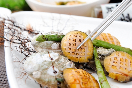 chopsticks grabbing grilled butter abalones with grilled asparagus, from white plateの写真素材
