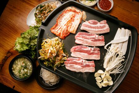 Raw pork belly with kimchi and seasoned green onion being grilled on grill with side dishの写真素材