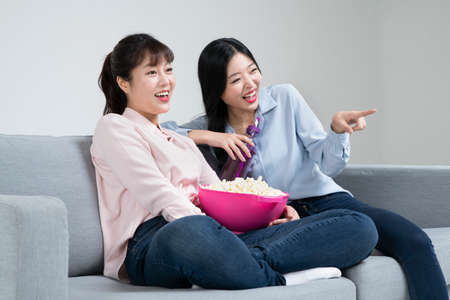 two Asian sisters, women with various facial expressions on sofa eating popcorn watching television
