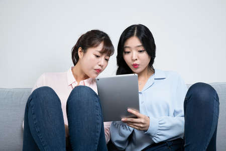 two Asian sisters, women on sofa watching video in digital tabletの写真素材
