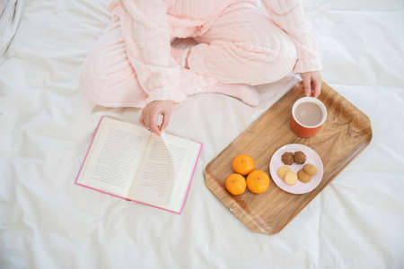 Asian woman with hair bun in pink pajamas on bed reading book and drinking coffee with tangerine and cookiesの写真素材