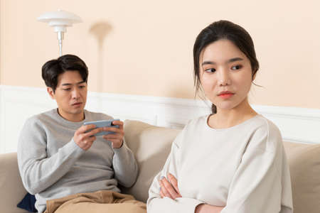 Asian wife angry at at husband using smart phone on sofa