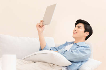young Asian man using tablet pc while lying on sofa