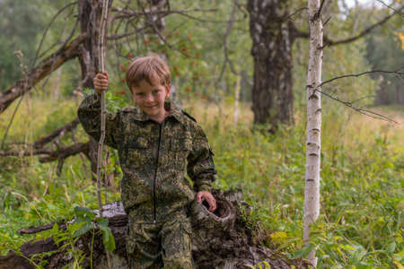 The boy in the autumn forest camouflage with a stick in his handの写真素材