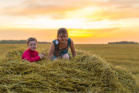 Boy and girl sitting on a stack of straw yellow smiling on the background of the setting, the rising sun in the cloudsの写真素材