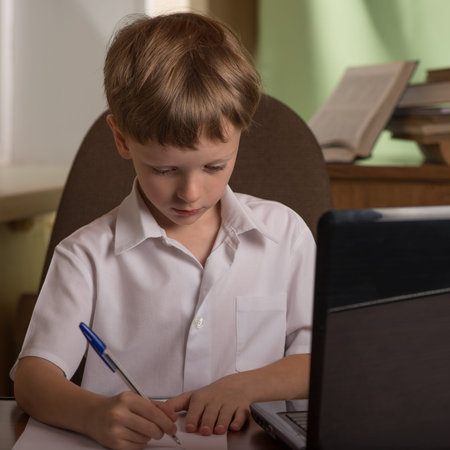 boy with laptop at the table taking notes with a pen on a sheet of paper
