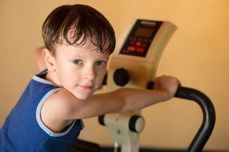 The child is trained on a stationary bike . Leads a healthy lifestyle. The boy develops muscles . Tired and sweaty . Sport generation.の写真素材
