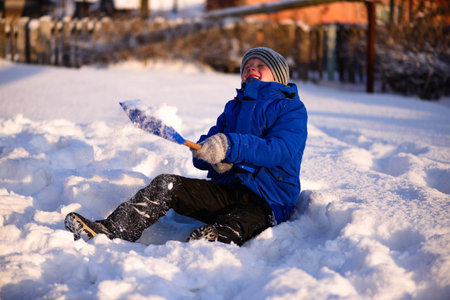 Guy in winter special clothes on a snow-covered field against the backdrop of a countryside landscape.Sits in the snow with a snowy children's spatula in her hands.の写真素材