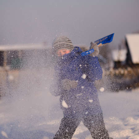 Guy in winter special clothes on a snow-covered field against the backdrop of a countryside landscape.He throws snow into the air with a snowy child's shovel.の写真素材