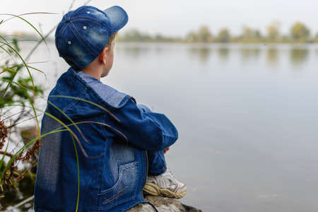 The child sits on the bank of the river on a rock and looking at the water away.Grabbing his knees with his hands.の写真素材