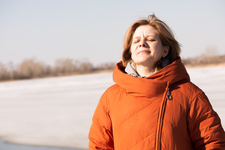 A woman in winter on the river bank without a headdress. Bright sun on a frosty day. Spring mood. Thaw and water without ice. Orange clothes. Orange shade. A comfortable day.の写真素材