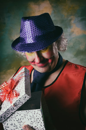 A man in a clown costume. A blue hat on his head, and a tie around his neck, a red vest. Foolish facial expression. Painted nose and lips. Gift cardboard box. There is a bow on the lid. Fun and celebration.の写真素材