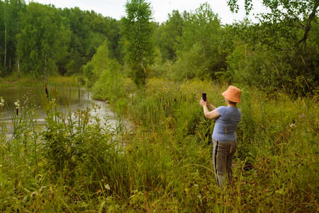 person is in nature near a reservoir. takes pictures, makes video broadcasts for social networks, records the environment. telephone is means of communication at a distance. world untouched by man.の写真素材