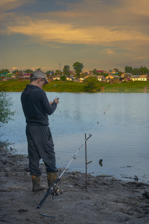 The guy at the reservoir is busy fishing. Consumed by attention to the fish biting, rewinds the fishing reel. Sits on a folding chair. Spends time outdoors. Keeping an eye on the fishing float.の写真素材