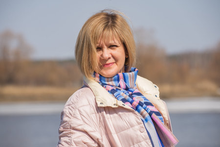 A woman stands on the bank of the river wearing a jacket and a scarf around her neck. Full face view. Short hairstyle blonde. Against the background of the shore of the reservoir. Native smile.の写真素材