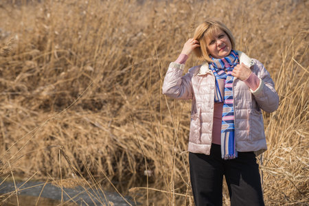 A woman stands on the bank of the river wearing a jacket and a scarf around her neck. Full face view. Short hairstyle blonde. Against the background of the shore of the reservoir. Native smile.の写真素材