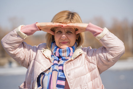 Stands on the bank of the river wearing a jacket and a scarf around her neck. Protected by hand from sunlight. Gaze is directed into the distance and into future. Against background shore reservoir.の写真素材