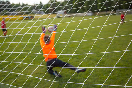 Goalkeeper catches the ball. Goal net in focus. Sports competition. Children's football on the field. Physical development. Children's sport. Sleight of hand. Ball Championship.の写真素材