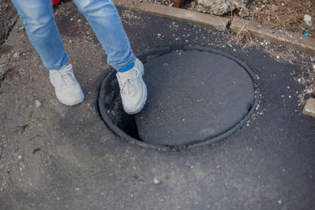 Open sewer manhole. Dangerous failure in the earth. Hole in the pavement. The girl steps into a dangerous place. Obstacle on the road. Failure in the ground. You can break a leg by stepping into a hole.の写真素材