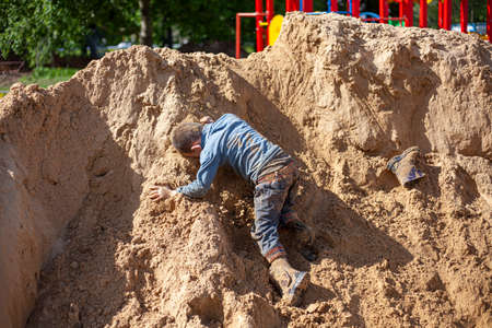 A boy falls from a mountain of sand. The child climbs up and breaks down. Children's game on the playground. Dangerous fall of the child. Active son shows his skills. Games for children without computers.の写真素材