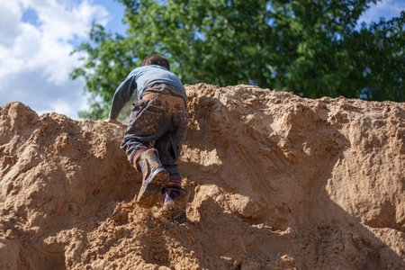 A boy falls from a mountain of sand. The child climbs up and breaks down. Children's game on the playground. Dangerous fall of the child. Active son shows his skills. Games for children without computers.の写真素材