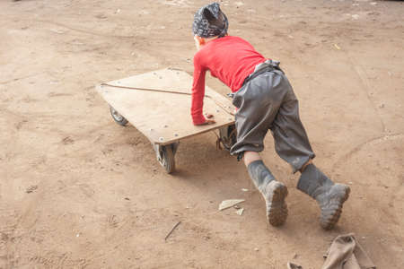 A boy plays with a cart on the street. A child in his fathers garage found amusement. Active child uses force. Energetic boy on a sunny day in the dust pushes a wheelbarrow. Children without gadgets.の写真素材