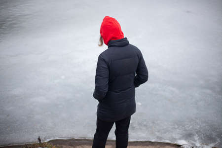 A lonely girl walks by the water. A teenager is tormented by loneliness. Woman in a red hood. Black clothes. Walk in the afternoon in the winter. The schoolgirl wandered into a deserted place.の写真素材