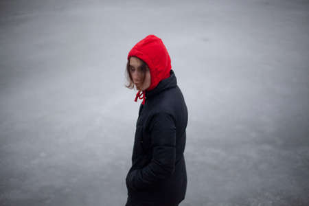 A lonely girl walks by the water. A teenager is tormented by loneliness. Woman in a red hood. Black clothes. Walk in the afternoon in the winter. The schoolgirl wandered into a deserted place.の写真素材
