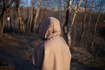 The girl walks in the evening in a warm jacket. Woman with a hood on her head. Unknown person in the park. Designer clothes of the original cut on the model. Warm coat for walking in cold weather.の写真素材