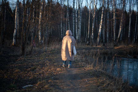 The girl walks in the evening in a warm jacket. Woman with a hood on her head. Unknown person in the park. Designer clothes of the original cut on the model. Warm coat for walking in cold weather.の写真素材