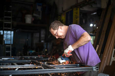 A man with a grinder in his hands cuts metal in his garage. A man with glasses does the work. Work during the holidays. Favorite business of an elderly person. Processing a metal profile to create a structure.の写真素材