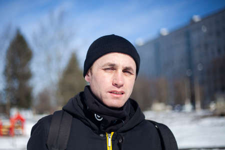 A man in a black hat and a black jacket. Portrait of a guy in winter on a sunny day in dark clothes. A young man is looking at the camera. Walk on a cold day.の写真素材