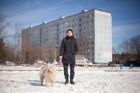 A guy in black clothes with a white dog. Dog walking in the winter. A young man is standing on the street. A modern guy holds a dog on a leash. Black suit for the cold season.の写真素材