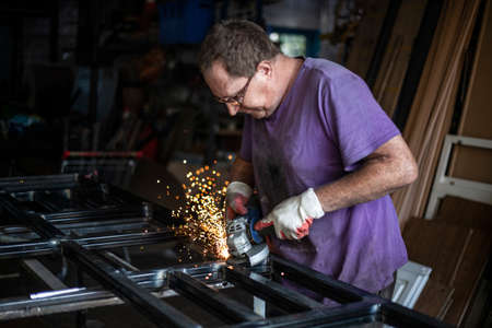 A man with a grinder in his hands cuts metal in his garage. A man with glasses does the work. Work during the holidays. Favorite business of an elderly person. Processing a metal profile to create a structure.の写真素材