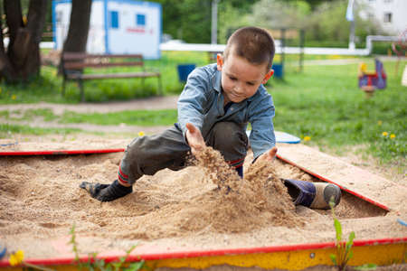 A child picks up sand. A boy plays in the sandbox. Walk with the child on a sunny day in the playground. The development of fine motor skills in a boy. Active lifestyle in children. Fine sand pours from hands.の写真素材