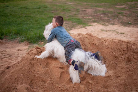 A child riding a white dog. A boy plays with a dog in the sandbox. A warm relationship between a child and an animal. Best friends. Playing on the playground with a dog. The son enjoys life. Rest without gadgets with a son.の写真素材