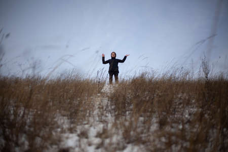 A girl stands in a field in winter. A young lady in black clothes is walking on the street. The model is removed in the open air. Teenage child. Overcast weather. Simple background with a man.の写真素材