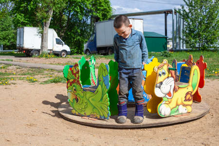 A child plays in the playground. Boy on the carousel. Active guy pushes a spinning platform. The joy of a child's walk. Active lifestyle. Healthy walk with the baby in the children's area on the street.の写真素材