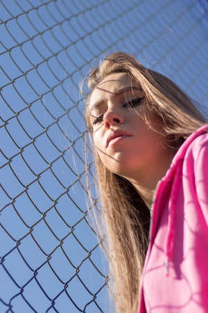 Girl near the trellised fence. Woman with long hair in a pink sweater. Sensual state of the model during shooting. Wrinkled face portrait. Beautiful makeup on the face. Mesh background when shooting.の写真素材