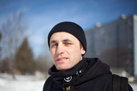 A man in a black hat and a black jacket. Portrait of a guy in winter on a sunny day in dark clothes. A young man is looking at the camera. Walk on a cold day.の写真素材