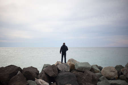 The guy walks along the promenade. A man by the sea. The guy is walking on large stones. The traveler came to the ocean. Seascape. Loneliness and adventure of a tourist. Rocky coast.の写真素材