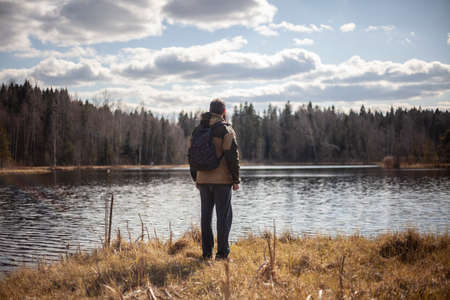 A man stands by the lake. The guy is dying on the water. Calm contemplation of nature. Spring walk in the forest. The young man froze in thought near a reservoir.の写真素材
