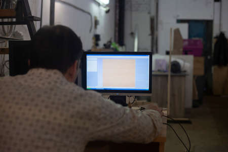 A man works at a laptop in a factory room. The guy runs the program. A shot from the back of an office worker. Doing small business. Screen brightness.の写真素材