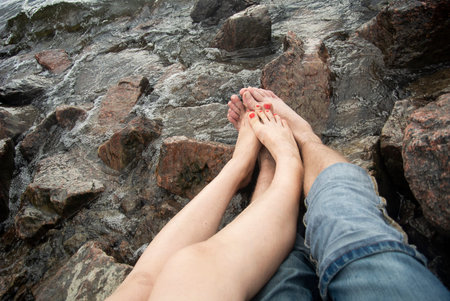 Legs of a man and a woman by the sea wave. Relax on a rocky beach. Fatigue of the legs after a walk. Foot care in the summer. Stones on the seashore. Romantic date on the beach.の写真素材