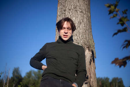 The guy is standing by a tree. A young man in a half-dress. A man in the woods. Outdoor recreation. The guy poses during the day.の写真素材